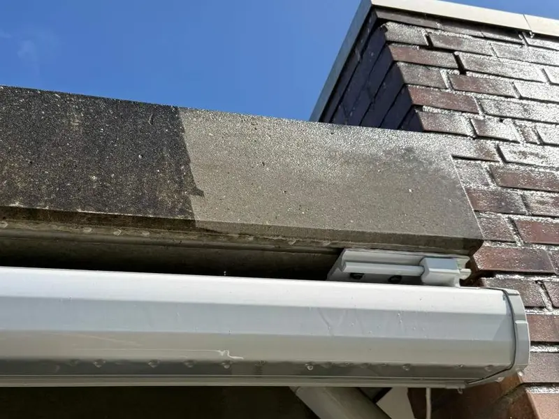 Betonnen dakrand boven een dakgoot: links nog donker van vuil, rechts lichtgrijs en schoon